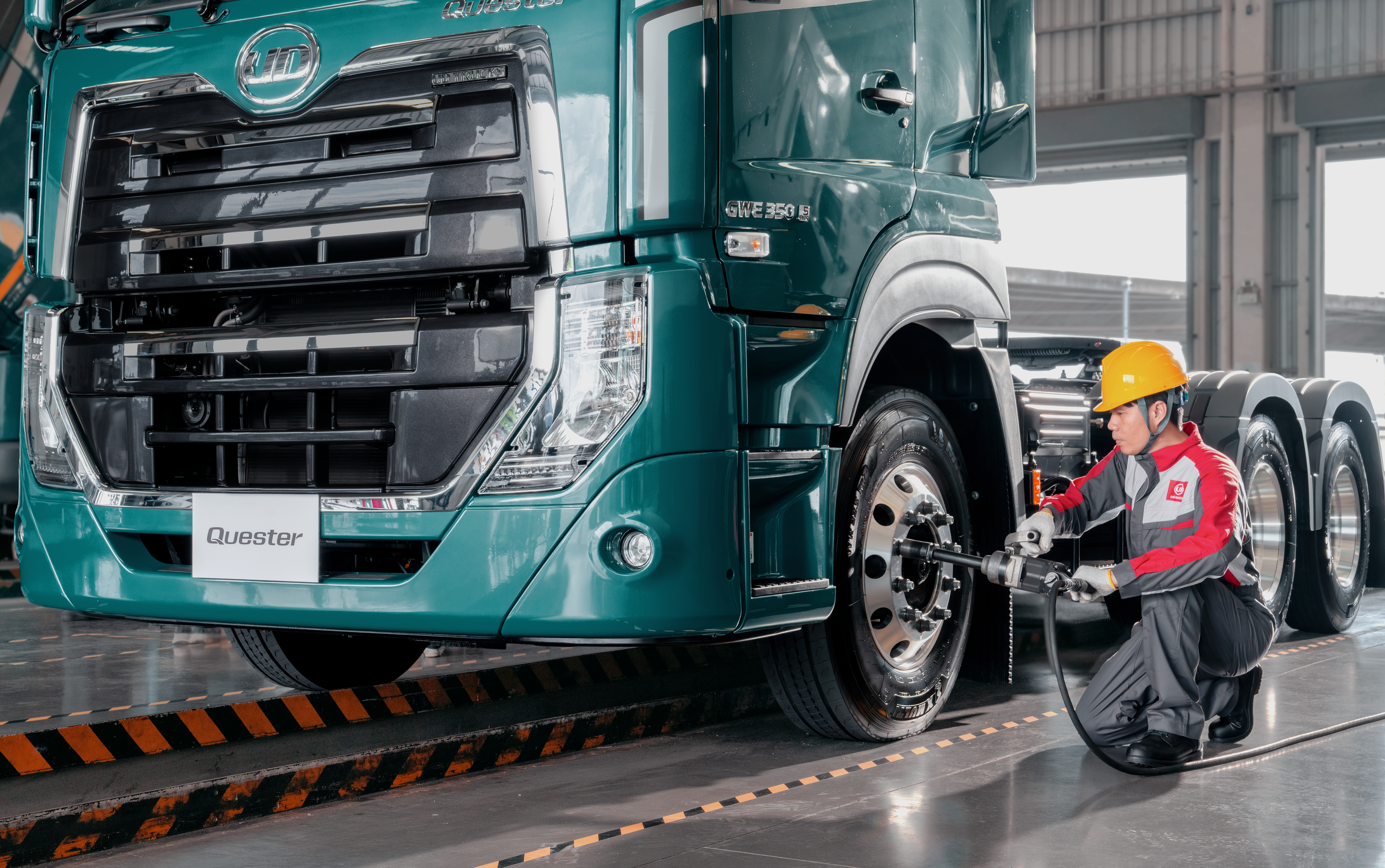 Quester Mechanic in safety gear checking a large green truck’s front wheel in a garage.