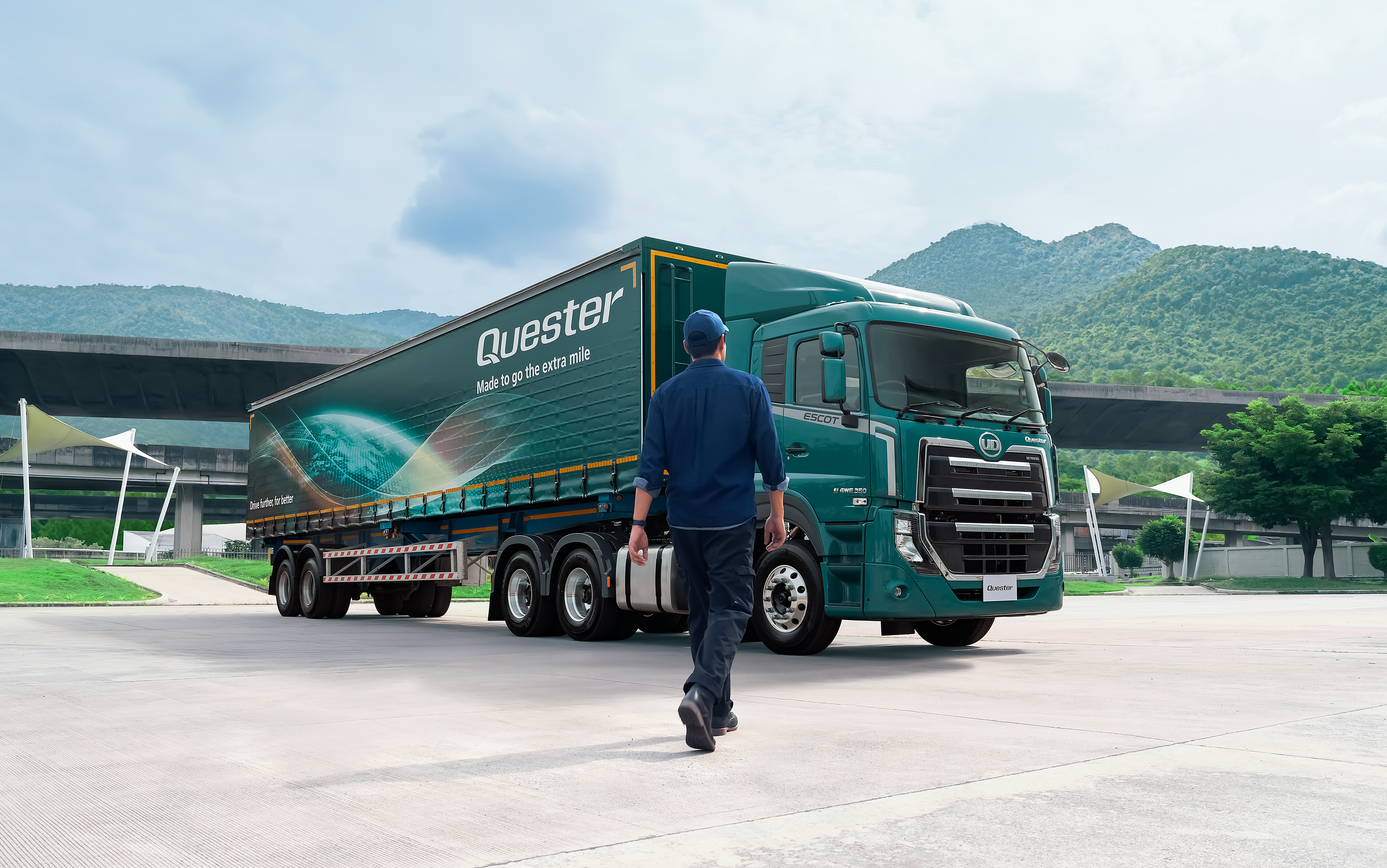 Quester Person walking toward a teal semi-truck in an outdoor lot with mountains in the background.