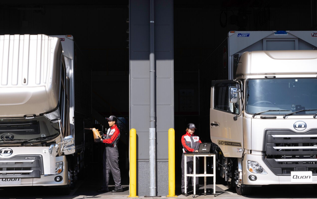 Two mechanics inspect large white semi-trucks parked in a service garage.