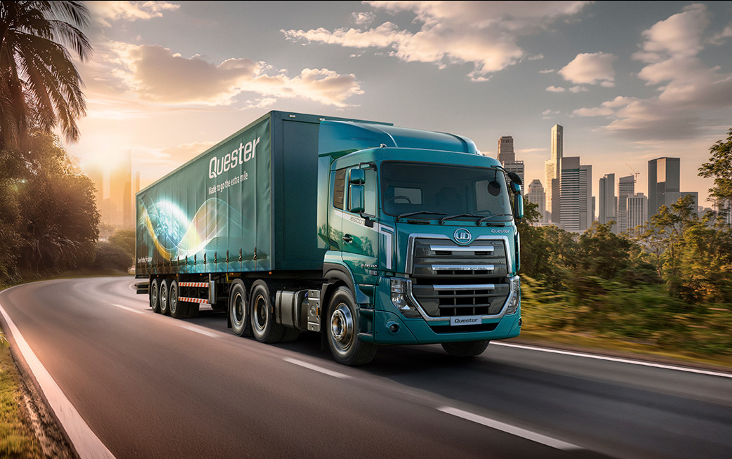Blue semi-truck driving on a road with city skyline in the background at sunset.