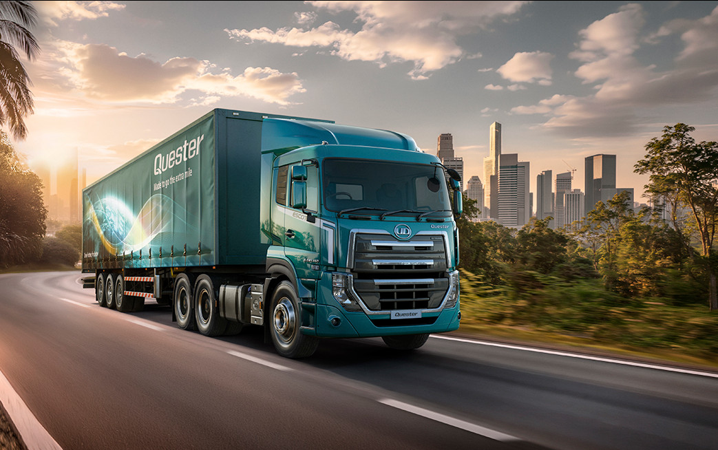 Blue semi-truck driving on highway with city skyline and palm trees at sunset.