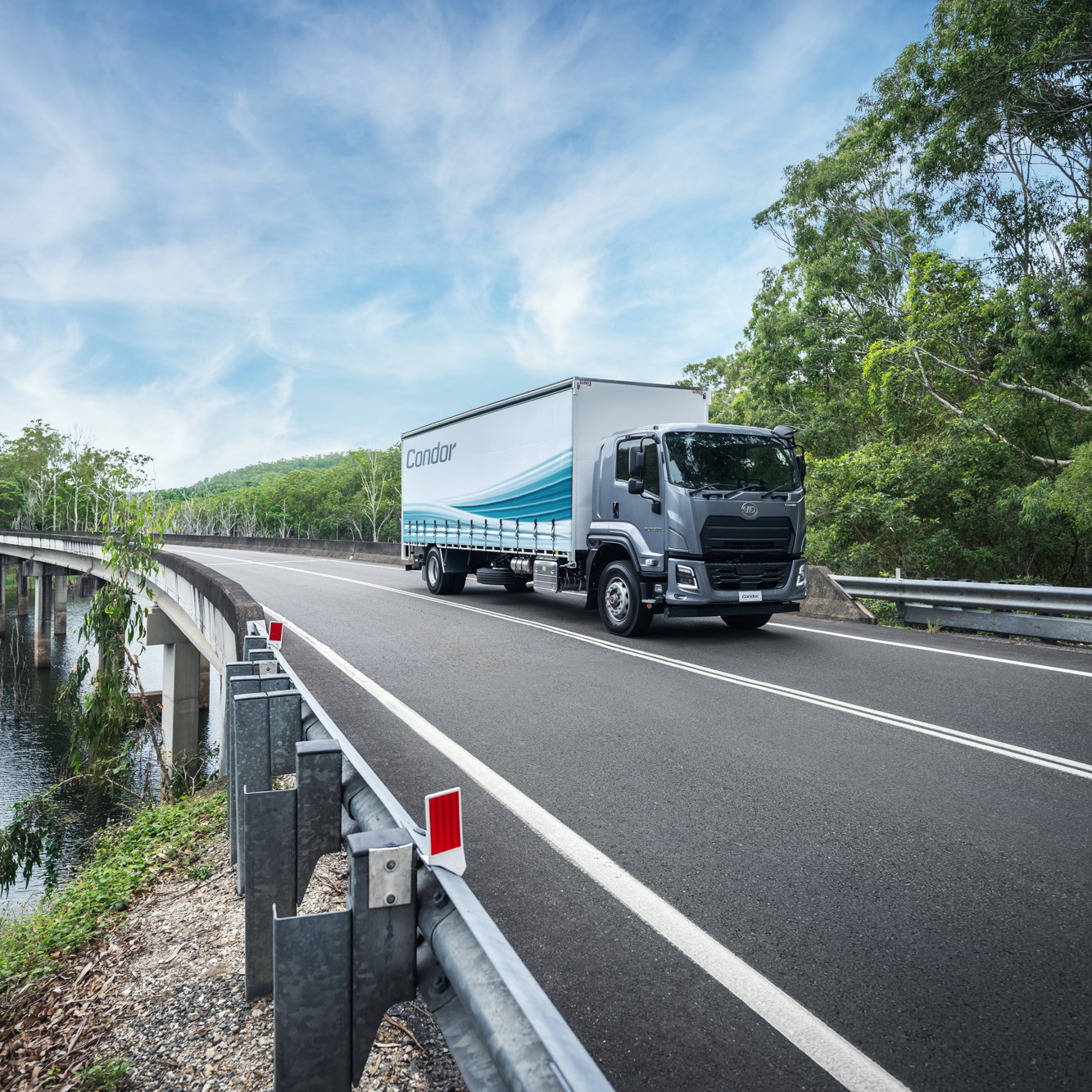 Gray delivery truck driving on a curved road surrounded by trees and blue sky.