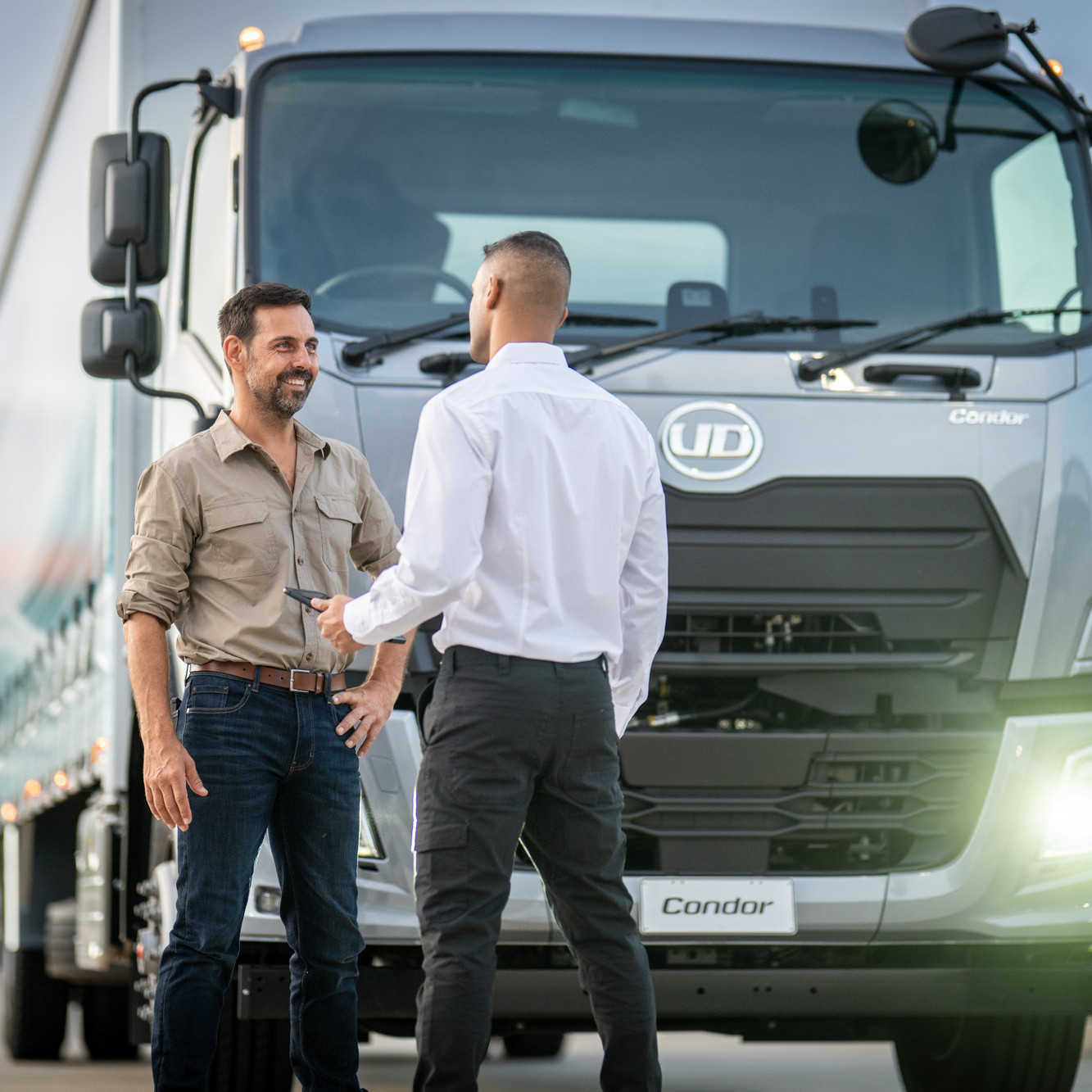 Two men talking in front of a large grey truck on a sunny day.