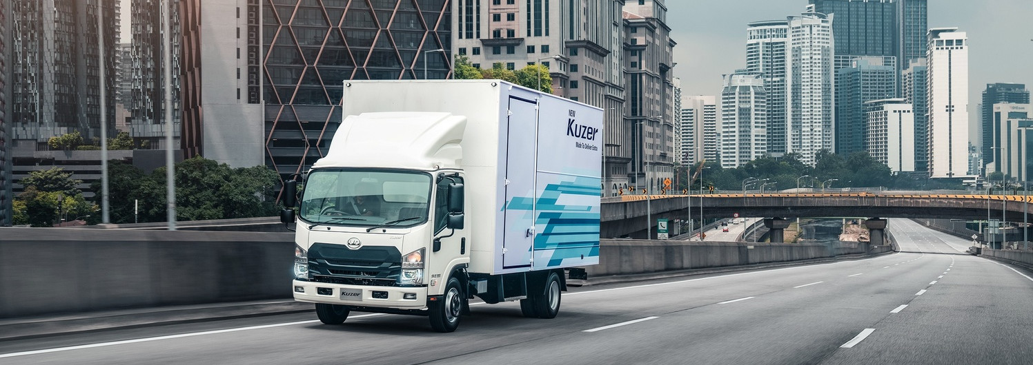 White delivery truck driving on empty city highway with tall buildings in background.