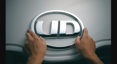 Hands holding a silver UD logo emblem on a gray surface.