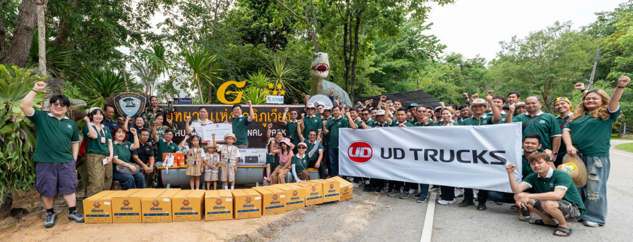 กลุ่มคนยืนถ่ายรูปกลางแจ้งถือป้าย UD TRUCKS ท่ามกลางต้นไม้และธรรมชาติ
