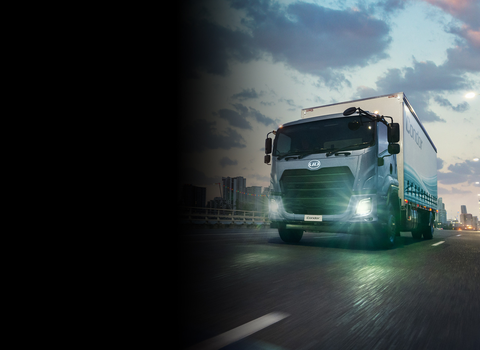 Large truck driving on city road at dusk with headlights on.