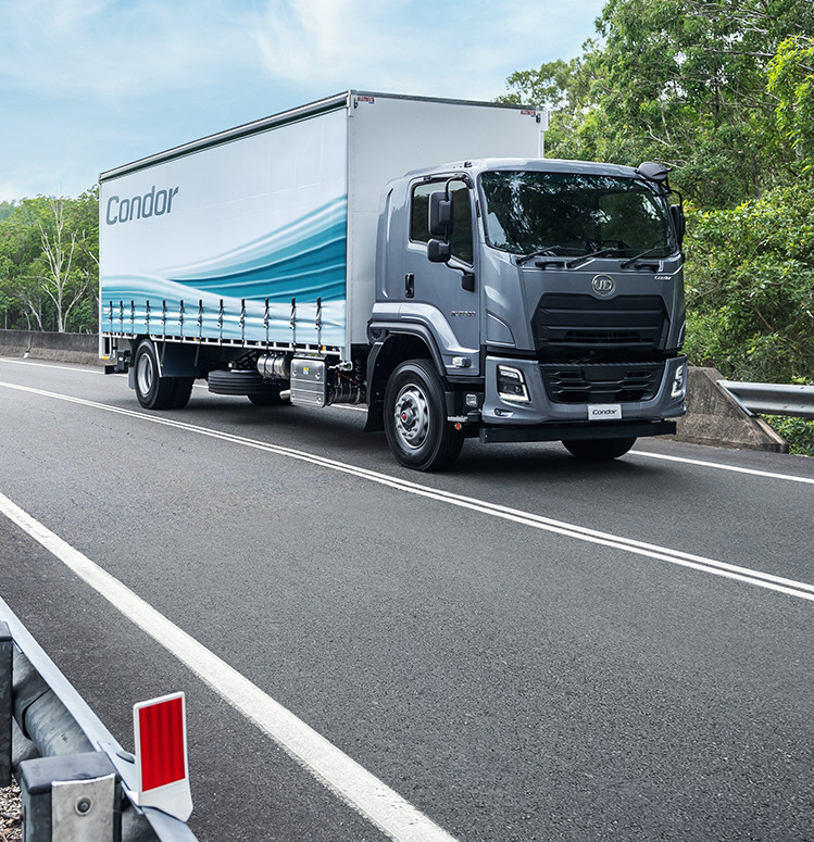 Large silver truck driving on an empty road surrounded by trees.