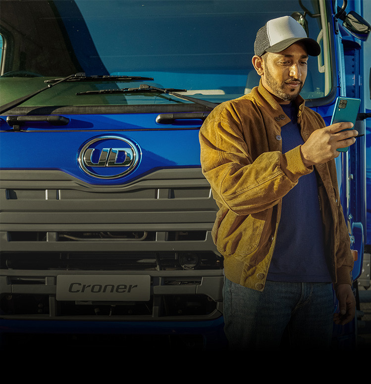 Lelaki memakai jaket coklat dan topi berdiri di depan lori biru, lihat telefon.